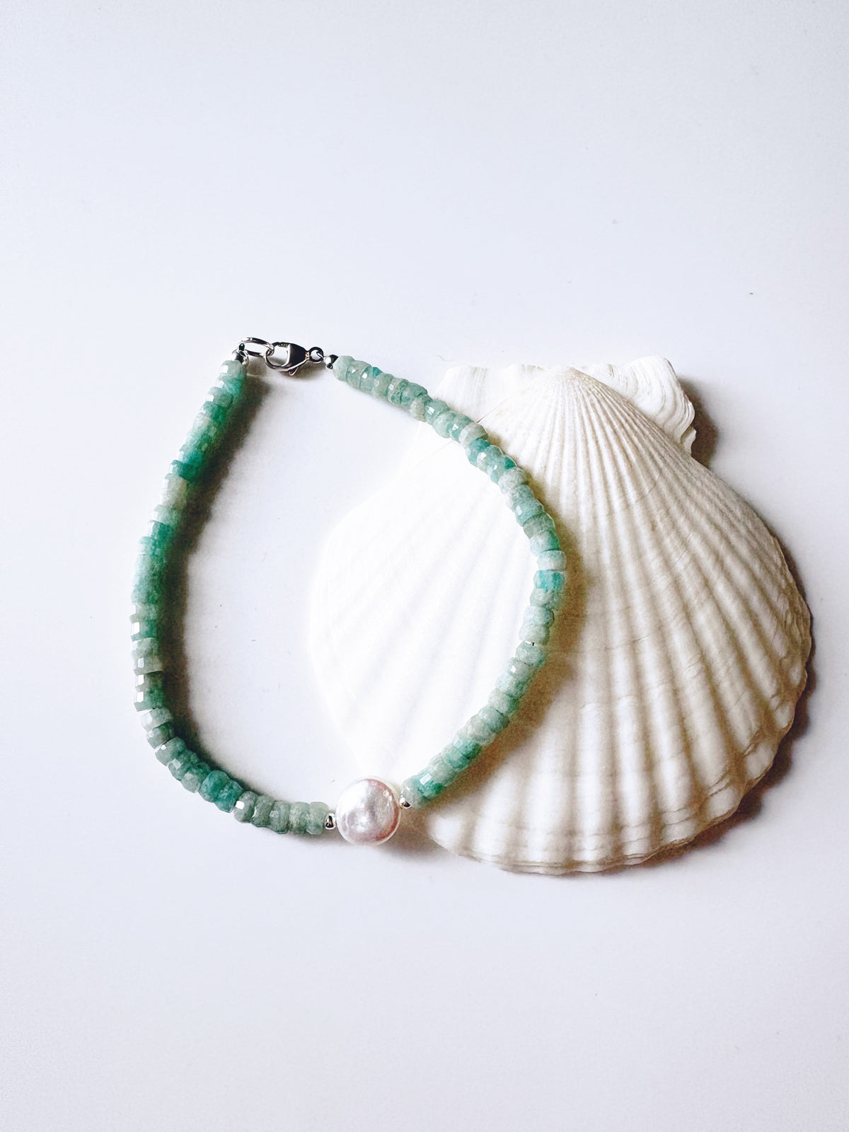 A beaded bracelet made of small green beads with a white pearl-like center charm and silver clasp, displayed on a white scallop shell against a light gray background.