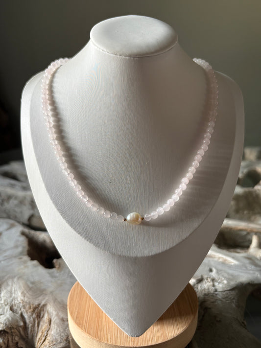 A delicate single-strand pearl necklace displayed on a white fabric jewelry bust, set against a rustic background of driftwood and a soft gray wall.