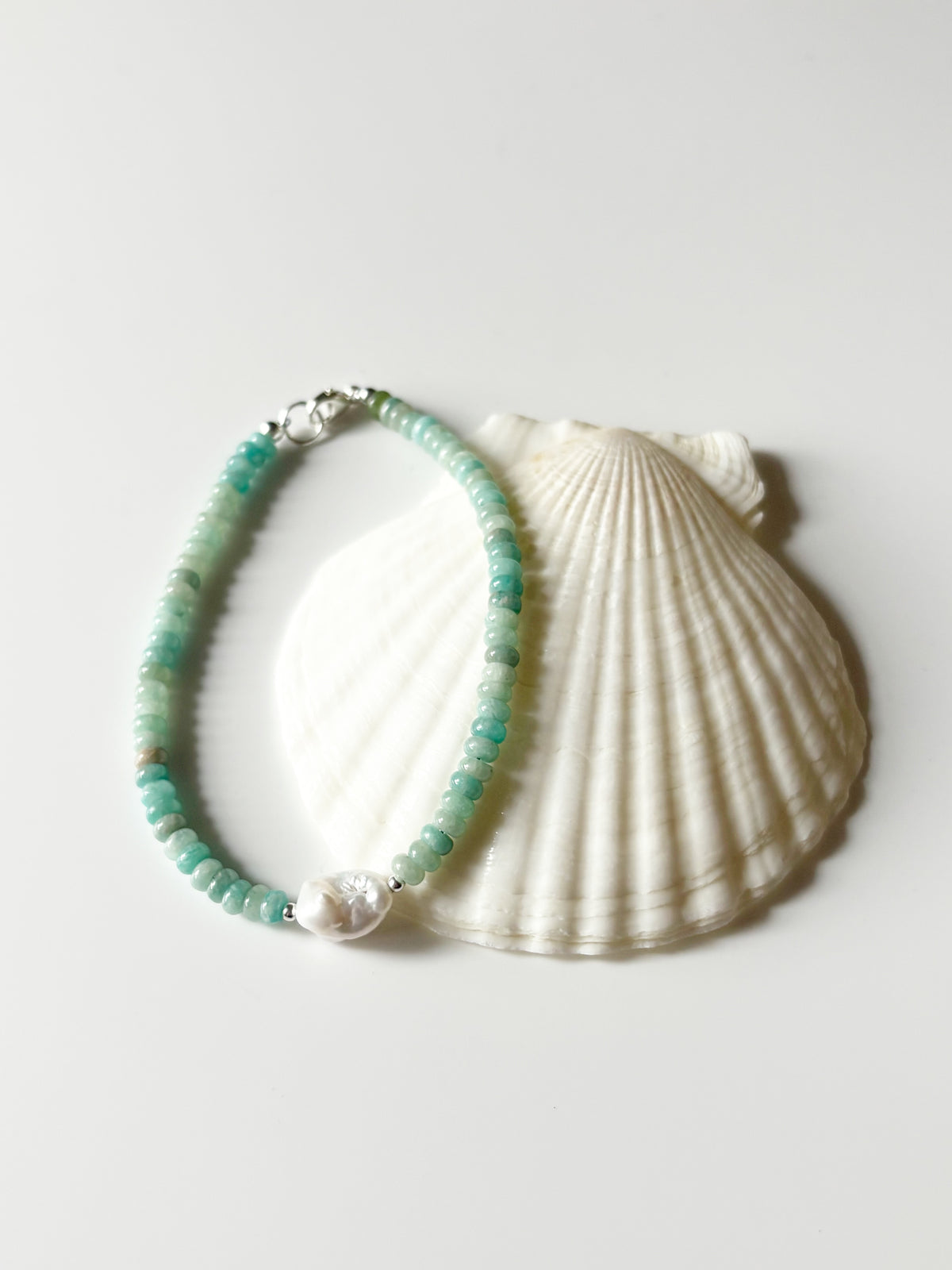 A beaded bracelet with light green and turquoise-blue beads, a white pearl accent, and silver-toned hardware, displayed on a white scallop shell against a light gray background.