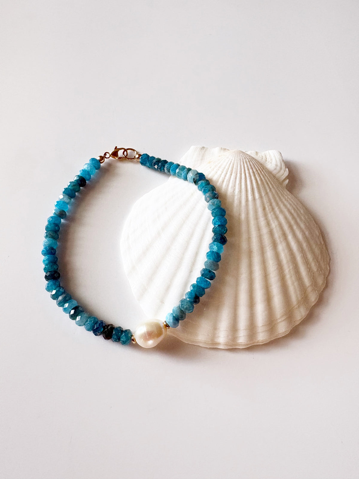 A beaded bracelet with turquoise-blue and dark blue faceted beads, a white pearl, and a gold clasp, displayed on a white seashell against a light gray background.