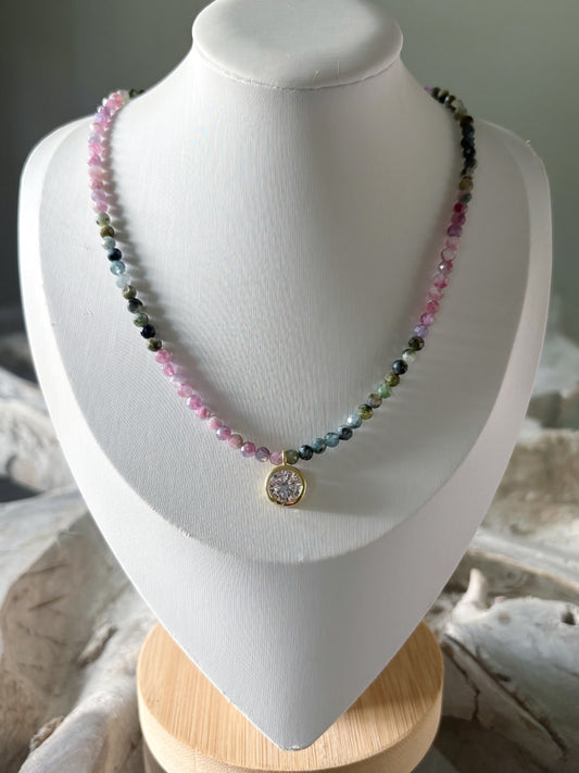 A beaded necklace displayed on a white jewelry bust. The necklace features small faceted beads in pink, green, blue, and black, with a gold-toned oval pendant containing a clear stone.