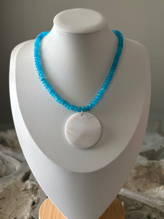 A bright blue beaded necklace with a large white circular pendant, displayed on a white jewelry bust.