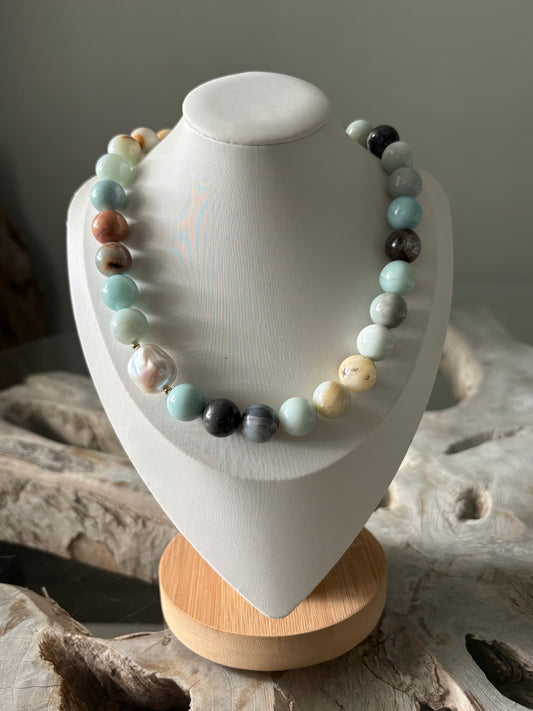 A beaded necklace made of round polished stones in pastel aqua, mint, cream, gray, brown, and black, displayed on a white jewelry bust with a light wood base.