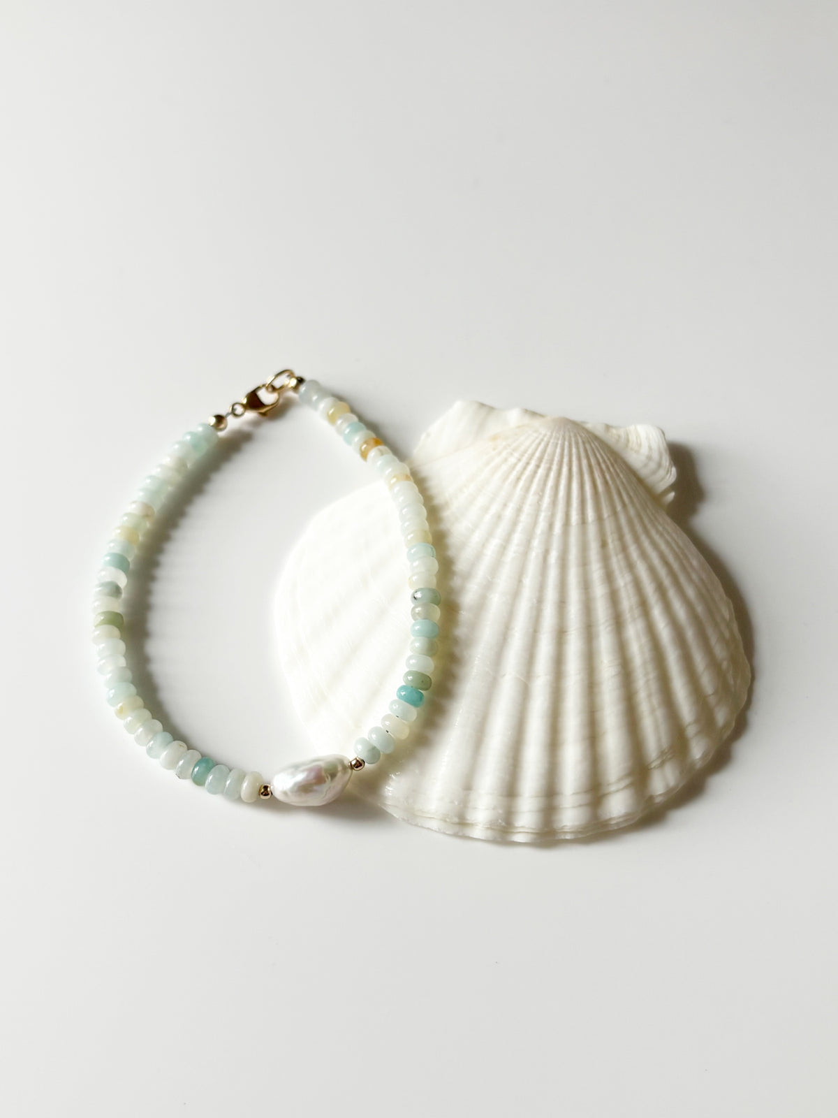 A beaded bracelet with light green and white beads, a white pearl accent, and gold-tone hardware, displayed next to a large white seashell on a light gray background.