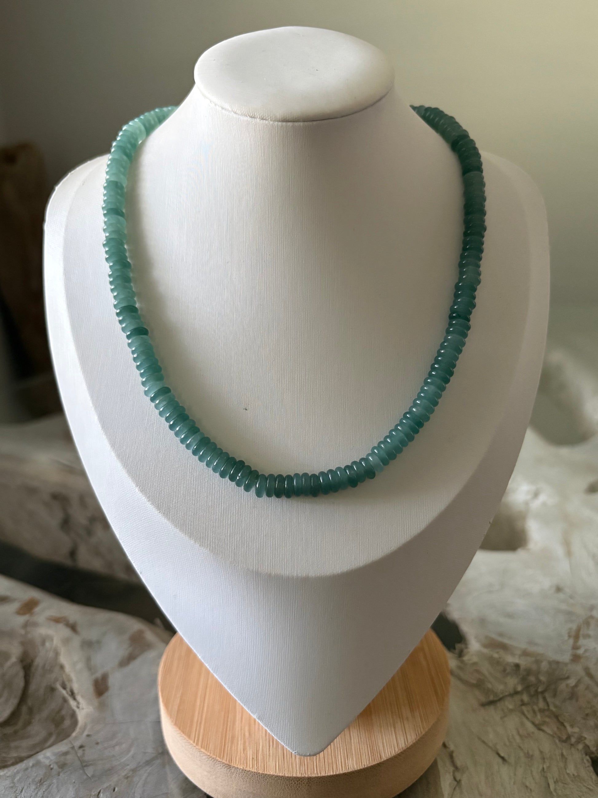 A chunky beaded necklace in a muted teal-green color, displayed on a white jewelry bust with a light wood base.