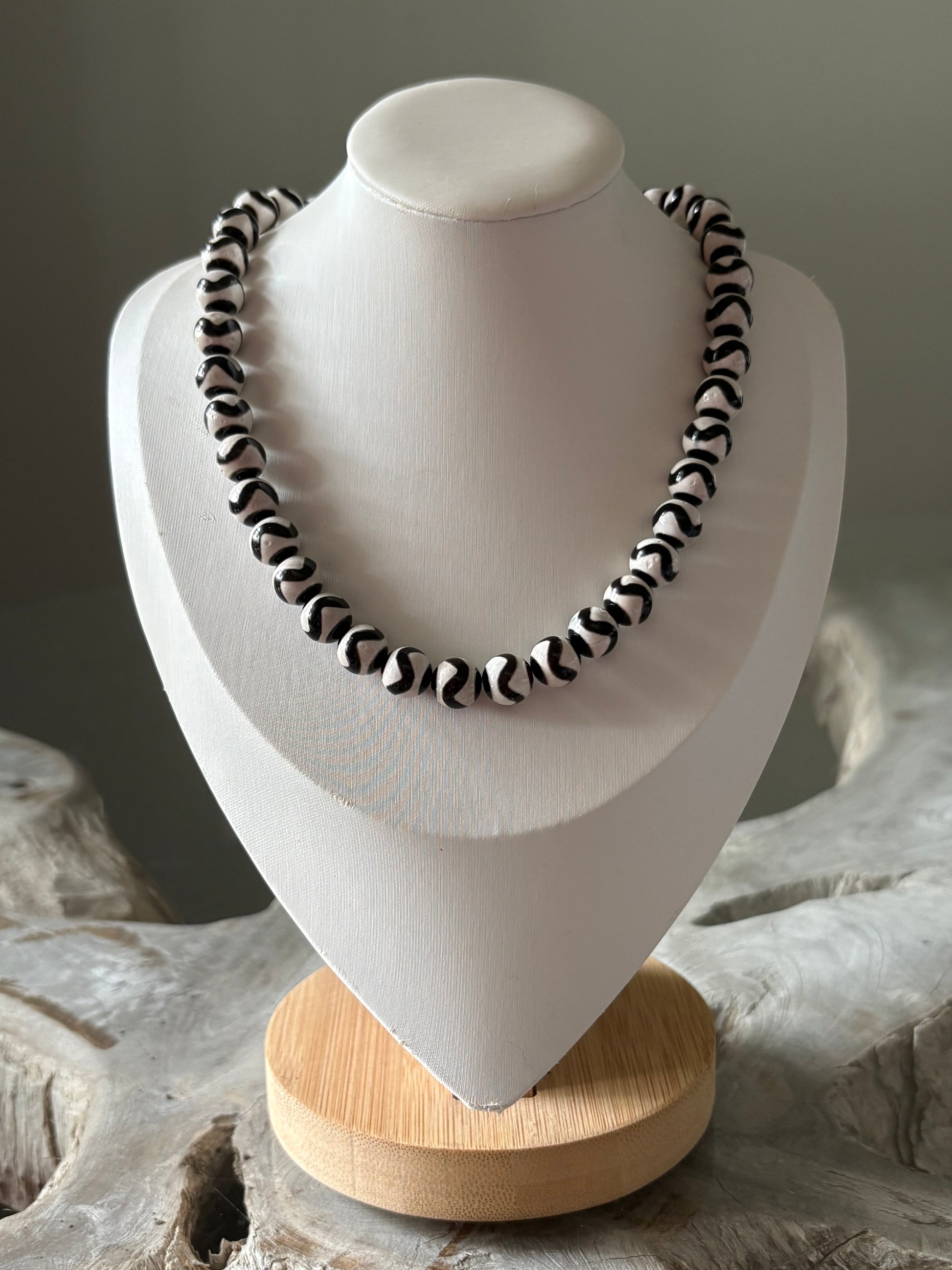 A black and white beaded necklace with a zebra-like pattern, displayed on a white jewelry bust with a light wood base.