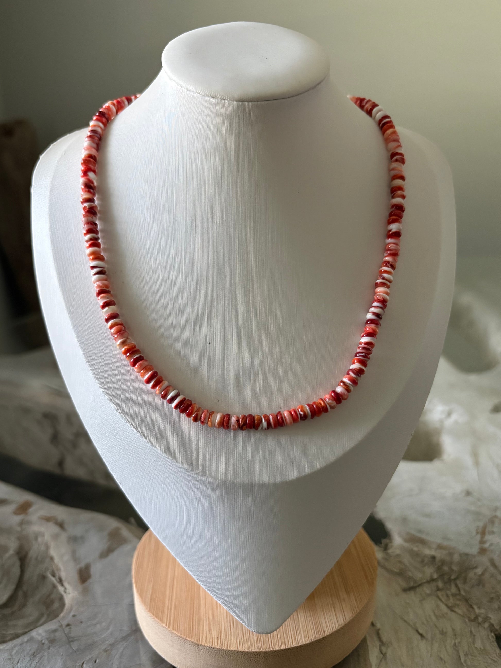 A beaded necklace made of small red and white beads, displayed on a white jewelry bust with a light wood base.