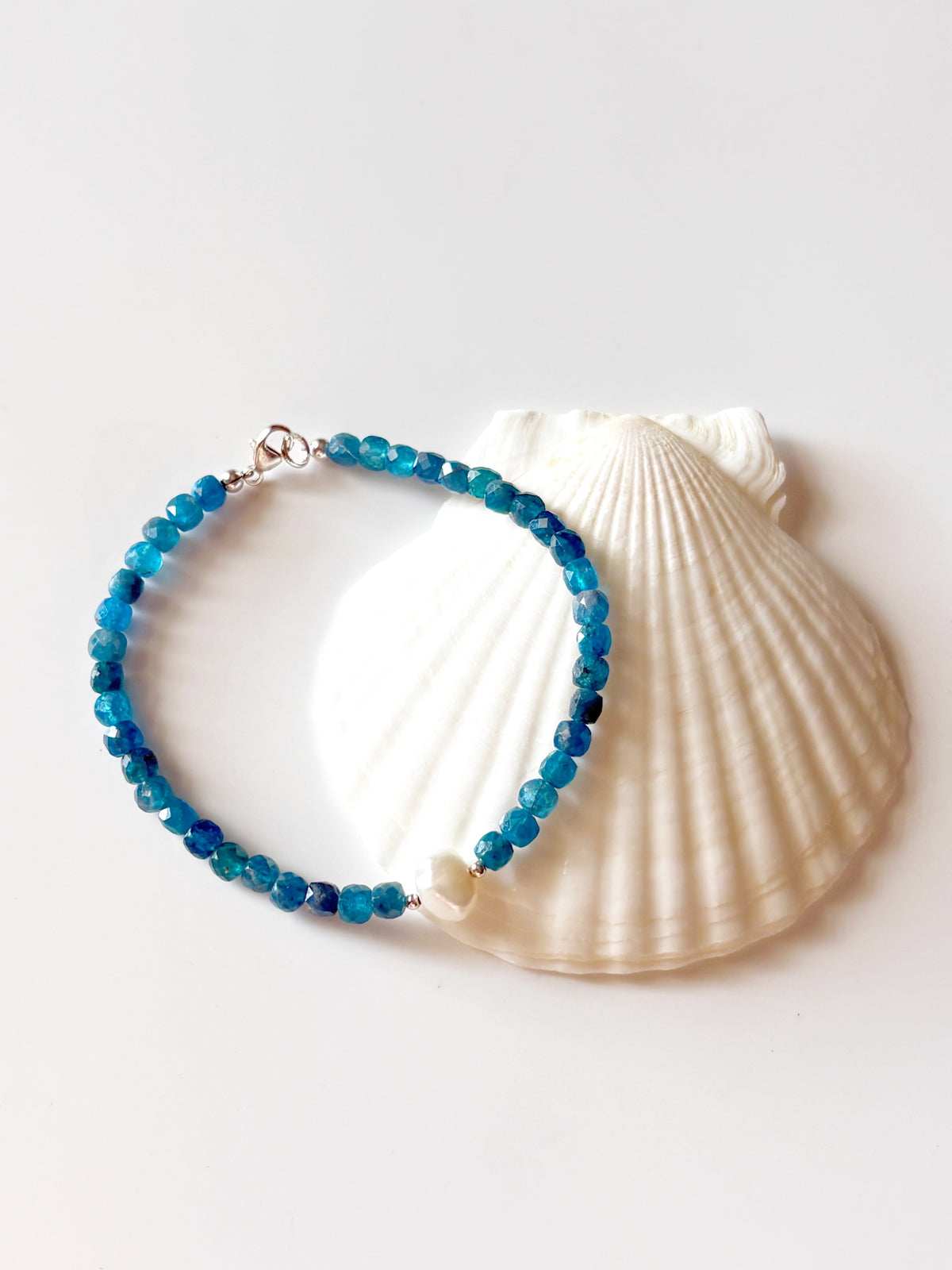 A beaded bracelet made of faceted blue and teal beads with a small white pearl accent and silver clasp, displayed on a large white scallop shell against a clean white background.