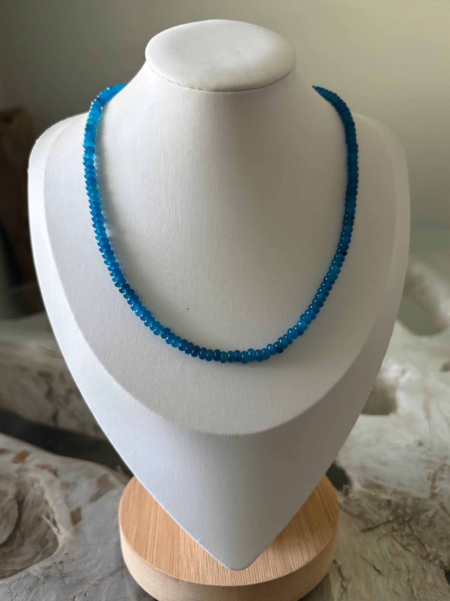 A beaded necklace in bright blue displayed on a white jewelry bust, with a light wood stand visible on the right side.