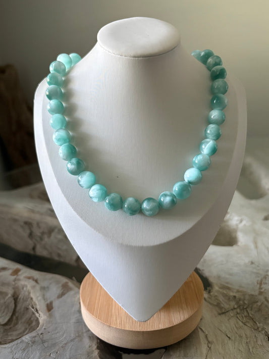 A light blue-green beaded necklace displayed on a white jewelry bust with a wooden base, set against a neutral stone background.