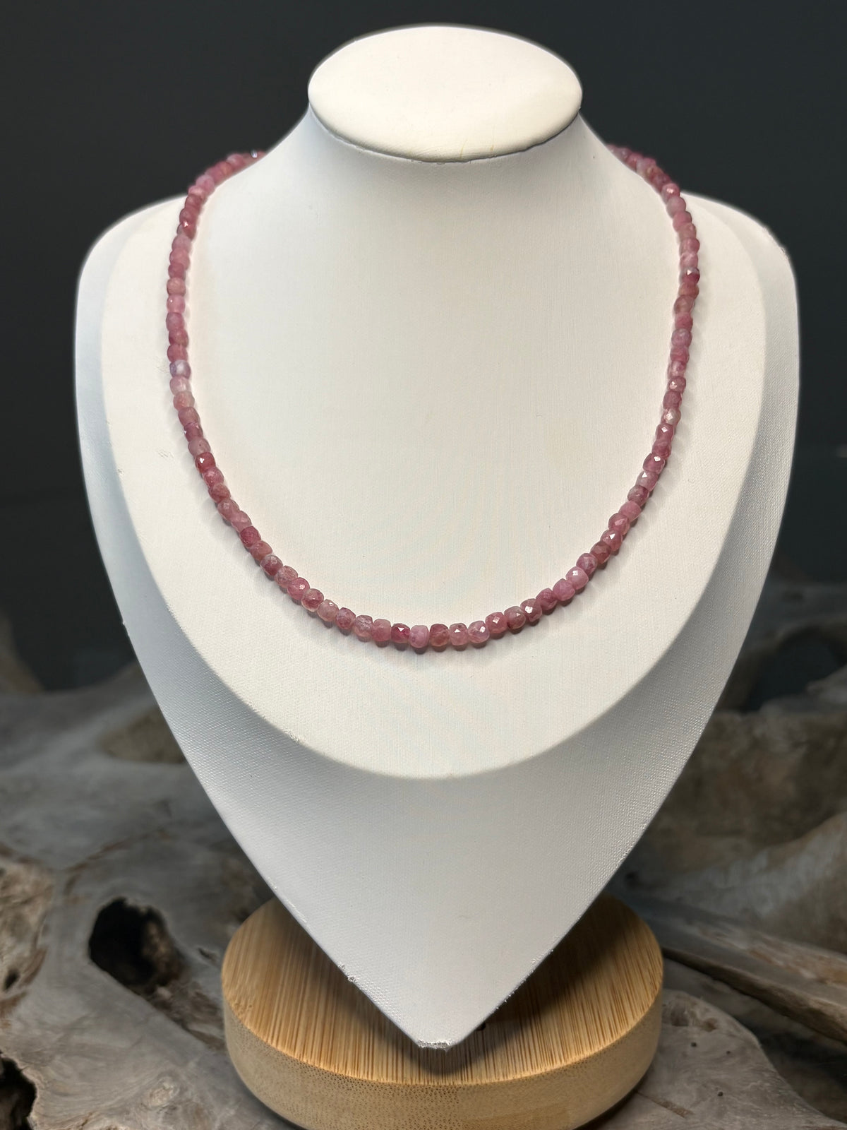 A pink beaded necklace displayed on a white jewelry bust with a wooden base, set against a dark gray background with natural wood props.