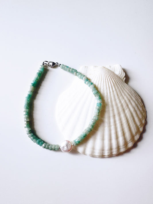 A beaded bracelet made of small green beads with a white pearl-like center charm and silver clasp, displayed on a white scallop shell against a light gray background.