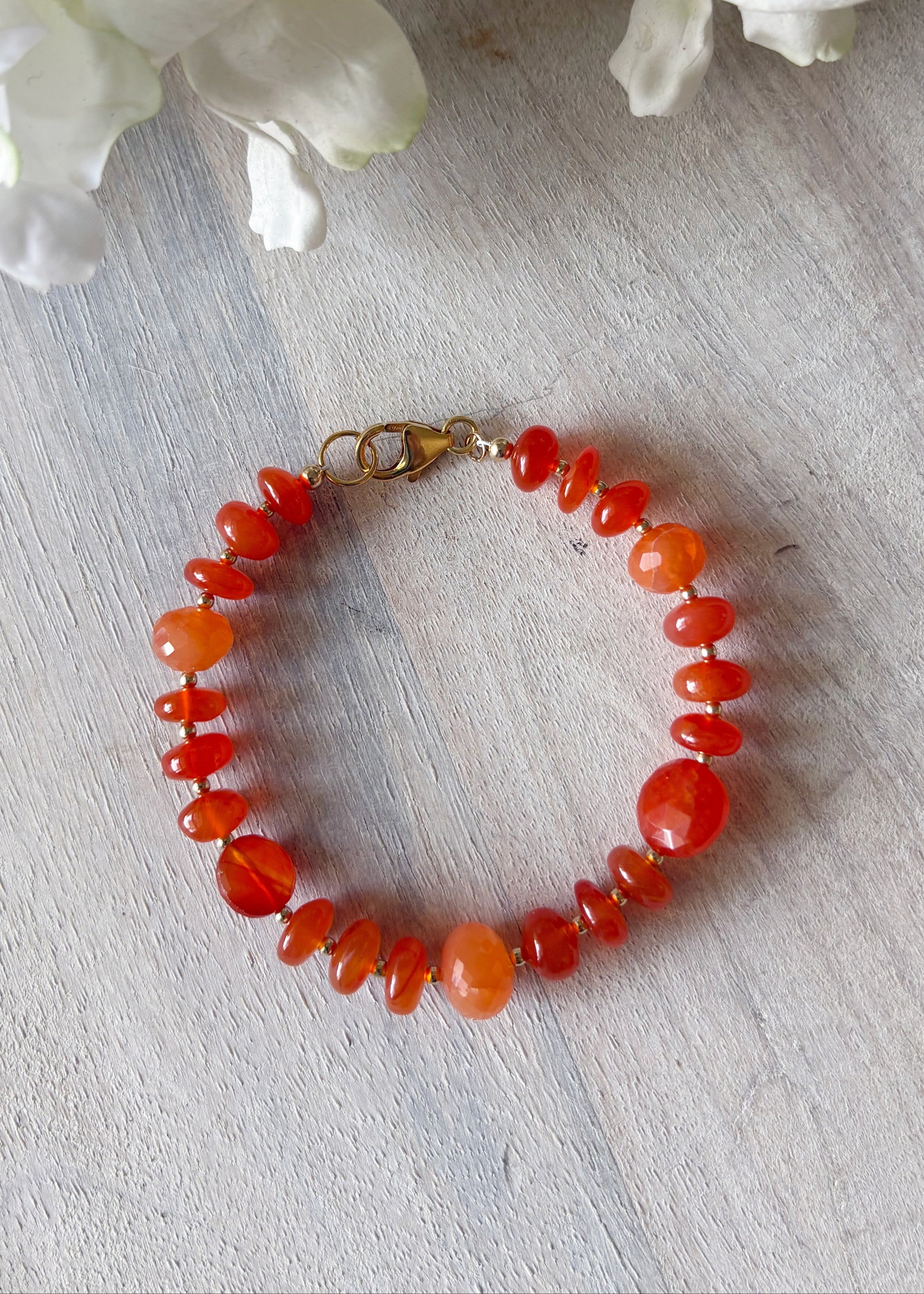 Orange beaded bracelet on a light wooden surface with white flowers in the background