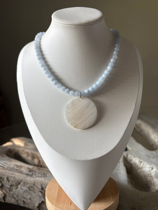 A light blue beaded necklace displayed on a white jewelry bust, with a matching round white pendant placed beside it. The bust is set against a softly blurred indoor background.
