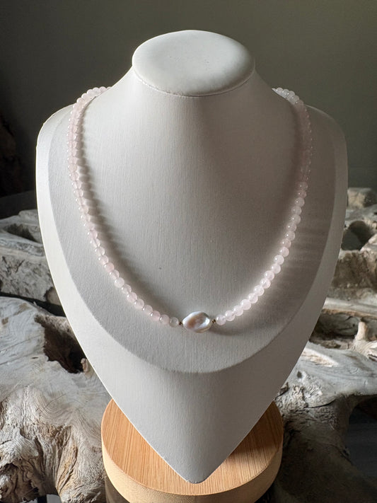 A single-strand pale pink beaded necklace displayed on a white jewelry bust, featuring a central white irregular shell bead, set against a rustic wood background.