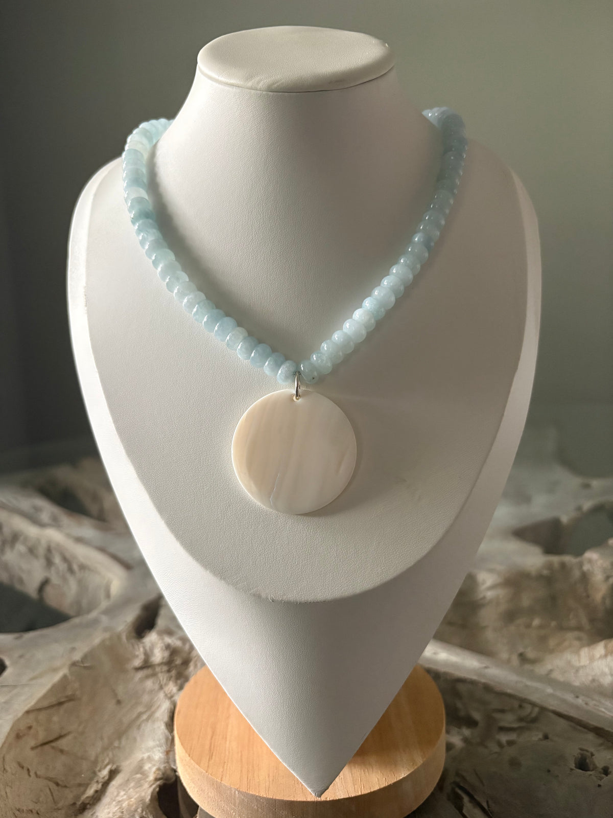A light blue beaded necklace with a round white shell pendant, displayed on a white jewelry bust.