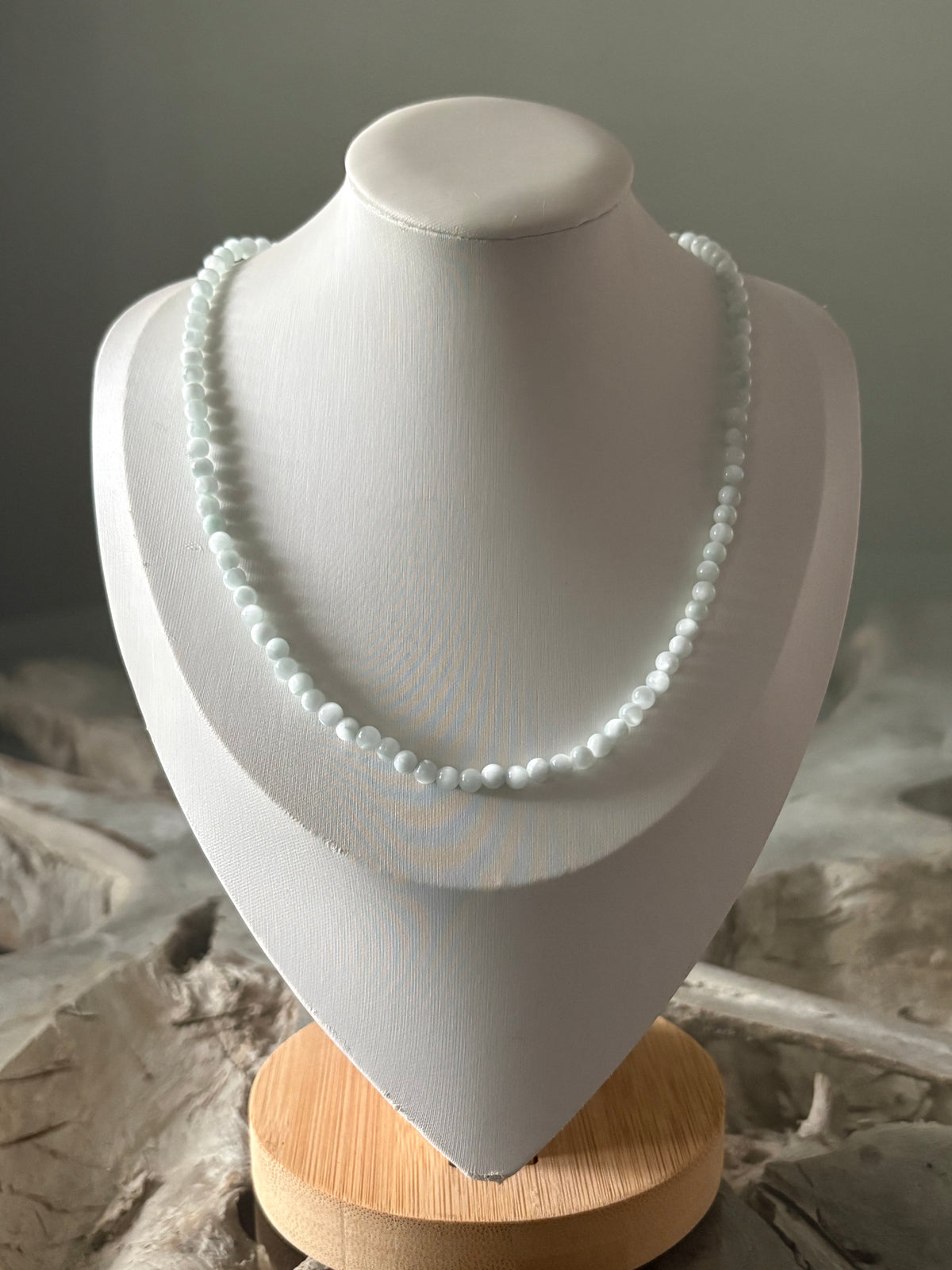 A delicate strand necklace of small, pale blue-green beads displayed on a white jewelry bust stand with a light wood base.